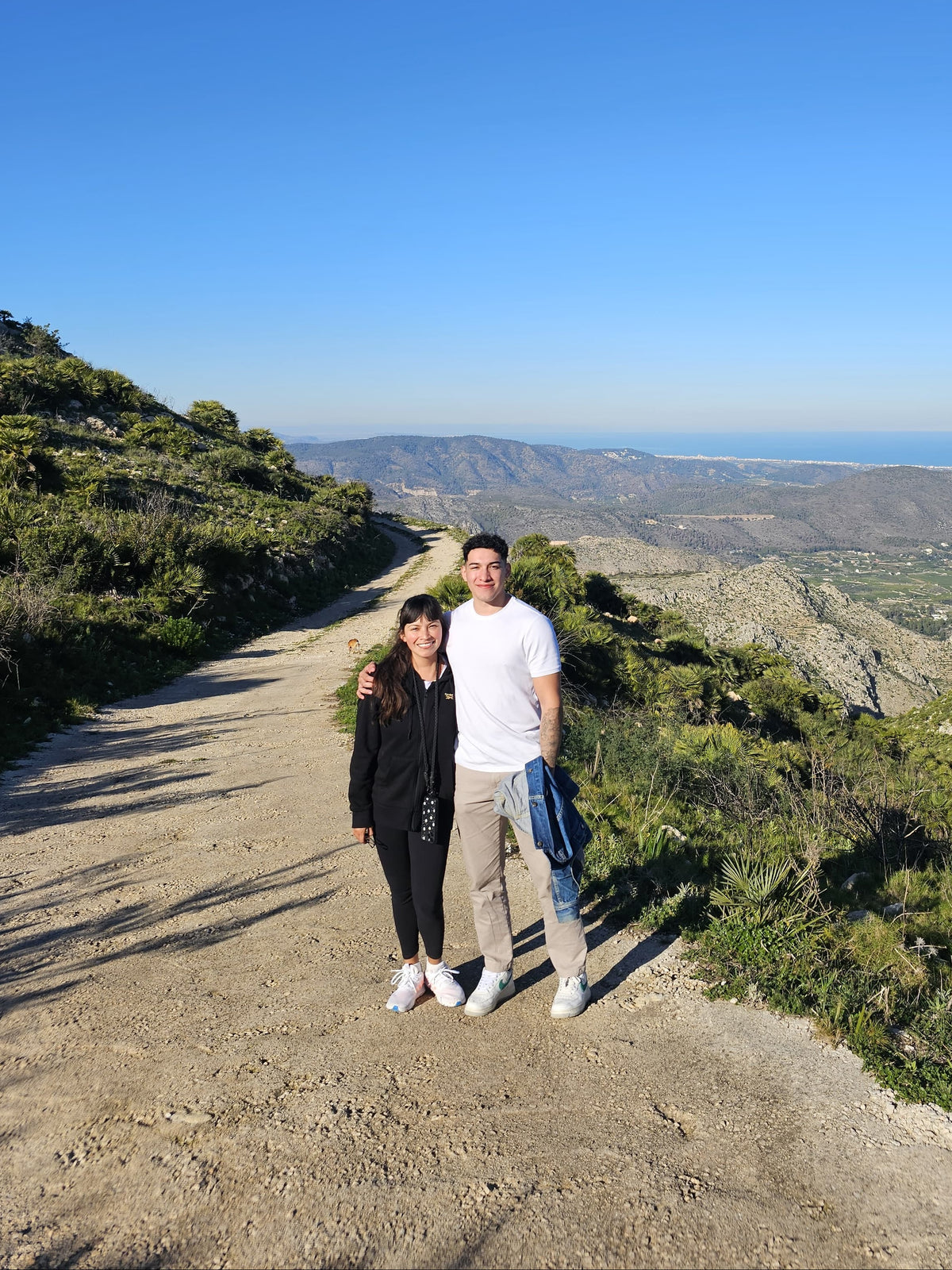 Two people standing on a scenic road with a clear blue sky and green landscape.