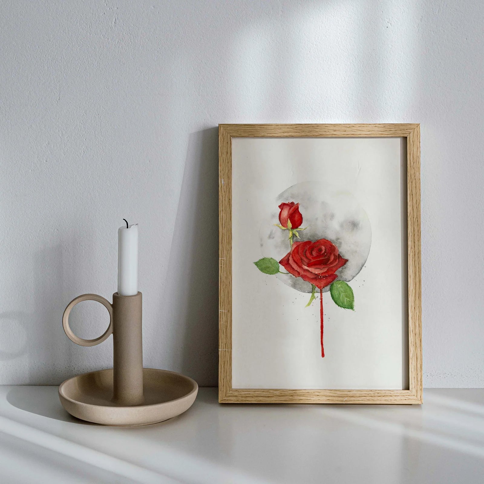 Framed artwork of a red rose on a white wall with a candle holder.